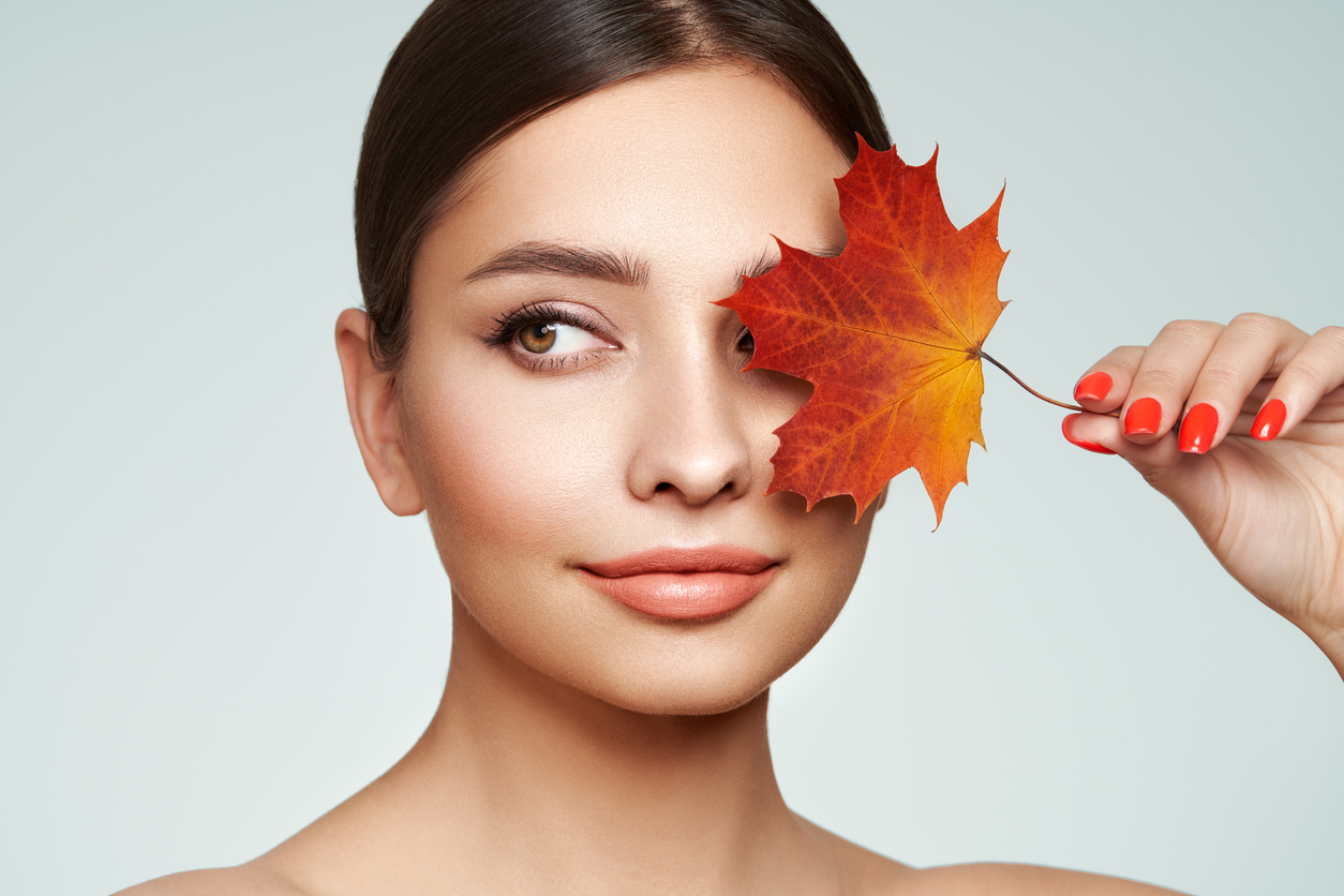 A gorgeous young brunette woman with red nails holds an orange & yellow leaf over her left eye