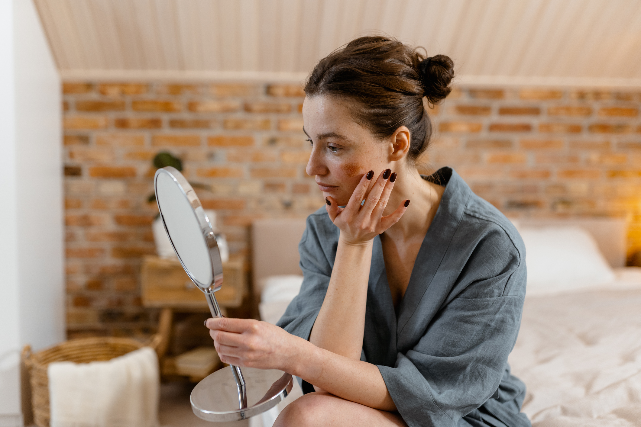 A woman looks at sun damage on her face in a mirror and considers going to an aesthetician.